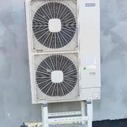 Zewnętrzna jednostka dwuwentylatorowej pompy ciepła Hitachi, zamontowana na białym stelażu na betonowym fundamencie przy ścianie budynku z widoczną izolacją fundamentu.