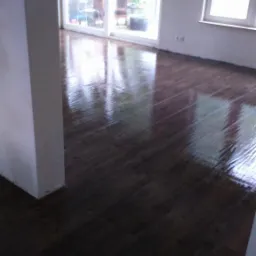 Wnętrze pokoju z drewnianą podłogą, dużym oknem balkonowym i oknem po prawej stronie, ściany pomalowane na biało, widoczny fragment kolumny.