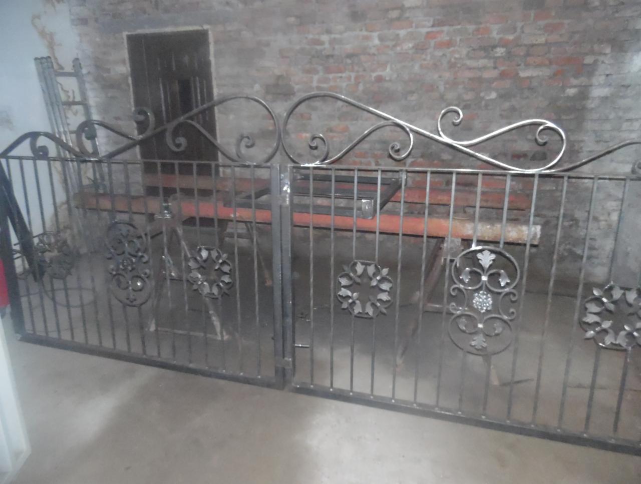 Wykute z metalu dwuskrzydłowe ogrodzenie z ozdobnymi elementami, w warsztacie na tle ceglanej ściany i drabiny.