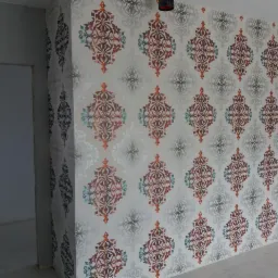 Fragment ściany pokrytej tapetą w ornamenty w odcieniach szarości, czerwieni i błękitu, widoczne fragmenty surowej ściany i ościeżnicy drzwiowej.