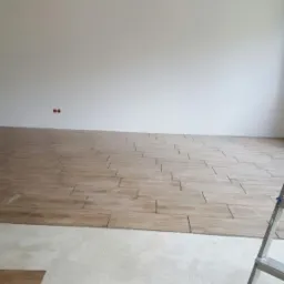 Wnętrze w trakcie układania podłogi z płytek imitujących drewno, widoczna drabina i surowy beton na części podłogi.