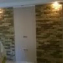 Fragment ściany z dekoracyjną okładziną imitującą kamień, oświetlonej od góry ciepłym światłem, z widocznym fragmentem białej ściany po lewej stronie.