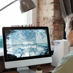 Kobieta przy biurku pracuje na komputerze, na ekranie widoczny napis 'Digital Marketing' na tle abstrakcyjnej grafiki miejskiej, obok żółty kubek i analiza danych na papierze.