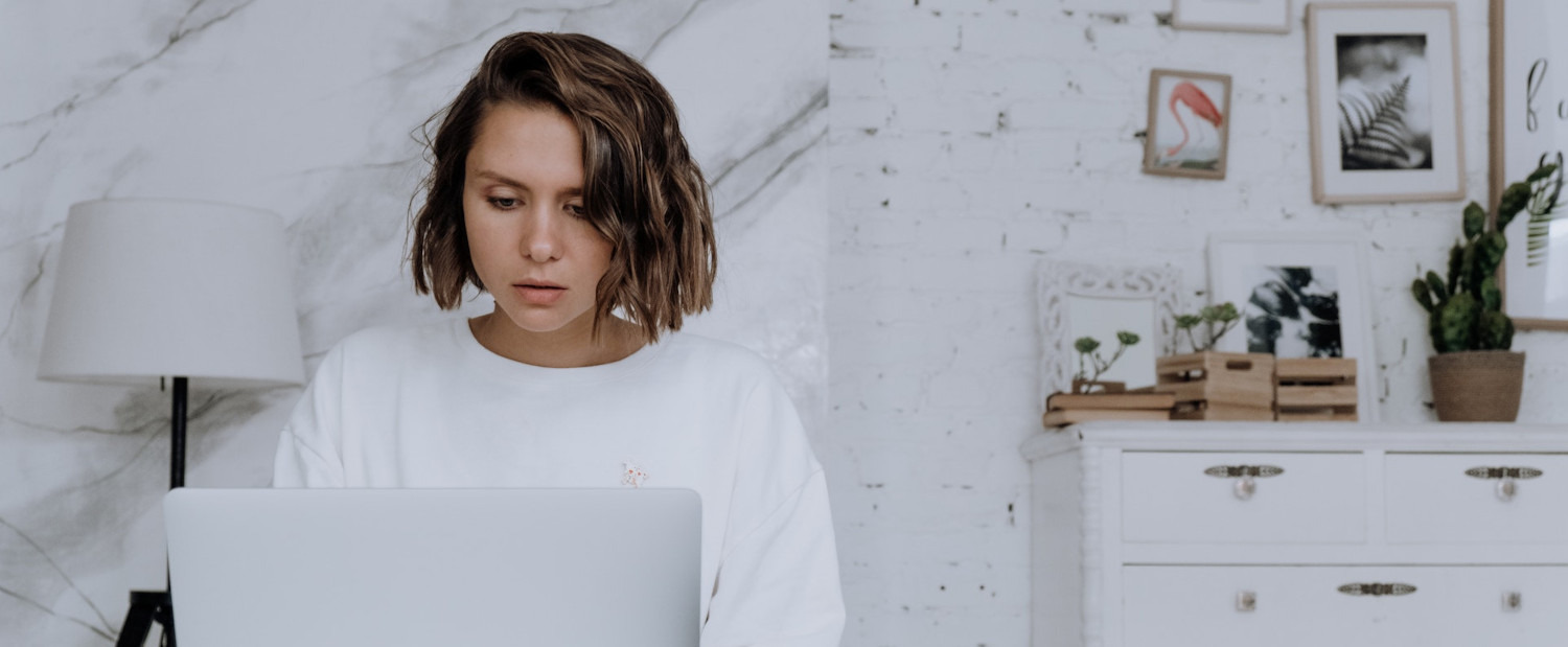 Kobieta w białej bluzie skupiona na laptopie, w tle biała ściana z dekoracjami i komoda