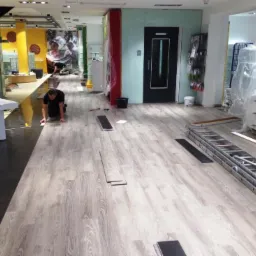 Pracownik na kolanach układa panele podłogowe o wzorze jasnego drewna w dużym pomieszczeniu, widoczna drabina, narzędzia i fragment czarnej folii podkładowej.