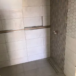 Wnętrze łazienki w trakcie prac wykończeniowych, widoczne ułożone płytki w różnych wzorach: prostokątne, heksagonalne i w romby, odpływ liniowy w podłodze, niezamaskowane rury doprowadzające wodę.