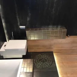 Nowoczesna łazienka z czarnymi, błyszczącymi płytkami ściennymi, drewnopodobną zabudową, podwieszaną toaletą i nietypową metalową kratką ściekową z napisem 'MIRAGE' w podłodze.