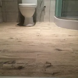 Jasnobrązowe panele podłogowe imitujące drewno w łazience z toaletą i kabiną prysznicową w tle.