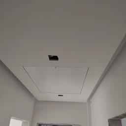 MG System - Sufit podwieszany z klapą rewizyjną i otworem na oświetlenie w trakcie prac wykończeniowych, widoczne niedoskonałości na ścianach i brakujące ościeżnice.