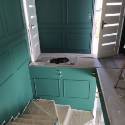 MG System - Malowanie klatki schodowej w Janikowie na kolor butelkowej zieleni z dekoracyjnymi panelami ściennymi, widoczne zabezpieczone schody i otwarte białe drzwi.