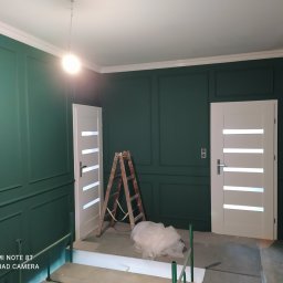 MG System - Wnętrze po malowaniu na ciemnozielono z dekoracyjnymi listwami ściennymi, białe drzwi z poziomymi przeszkleniami, drabina malarska i zabezpieczona folią podłoga przed dalszymi pracami.