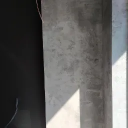 Ściana pokryta dekoracyjną, szarą strukturą z widocznymi nieregularnościami i zacienieniami, obok czarna ściana z wystającymi przewodami elektrycznymi.