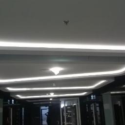 MG System - Sufit podwieszany z płyt karton gips z oświetleniem LED w kształcie prostokątów, widok z dołu na korytarz z przeszklonymi drzwiami.