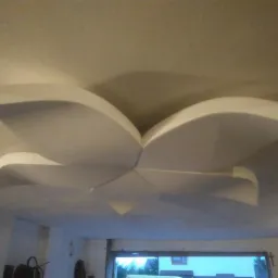 Dekoracyjny sufit podwieszany w kształcie kwiatu, wykonany z białych płyt, widoczny z dołu, z fragmentem okna w tle.