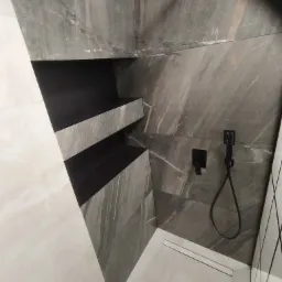 Nowoczesna kabina prysznicowa z szarymi płytkami imitującymi marmur, czarną armaturą i wnękami na kosmetyki wykończonymi w czerni.