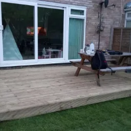 Drewniany taras przydomowy z widocznym stołem piknikowym, parasolem i elementami domu w tle.