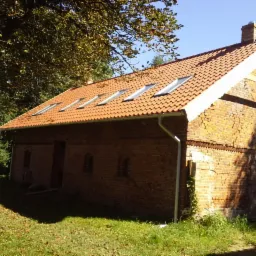 Stary, ceglany budynek gospodarczy z nową, czerwoną dachówką i czterema oknami dachowymi, widoczny komin i rynna, otoczony zielenią.