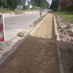 Przygotowanie podłoża z piasku i obrzeży betonowych pod chodnik lub drogę rowerową wzdłuż asfaltowej jezdni, widoczne bariery drogowe i budynki w tle.