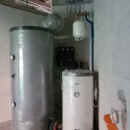 Dwa zbiorniki na wodę, jeden szary owinięty folią, drugi biały również owinięty folią, oba z manometrami, w surowym pomieszczeniu z betonową podłogą i instalacją rurową pod drewnianym stropem.