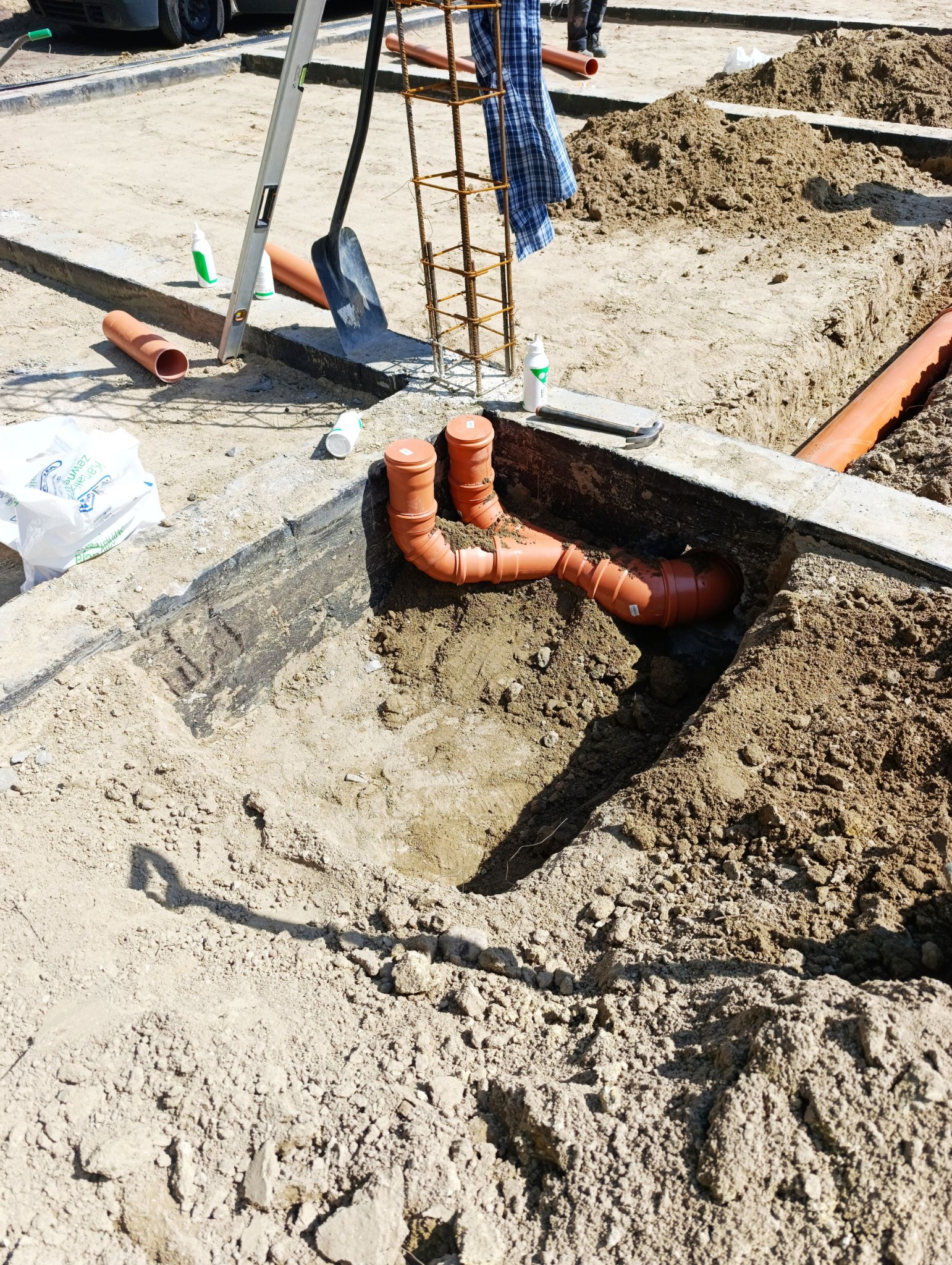 Wykonane przyłącze kanalizacyjne z pomarańczowych rur w wykopie, widoczne złącza kolankowe, ziemia i elementy szalunku.