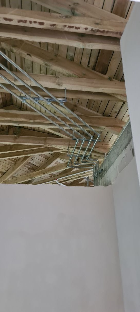 Szare rury instalacyjne zamocowane pod drewnianą konstrukcją dachu, widoczne elementy montażowe, ściana w tle w trakcie wykańczania.