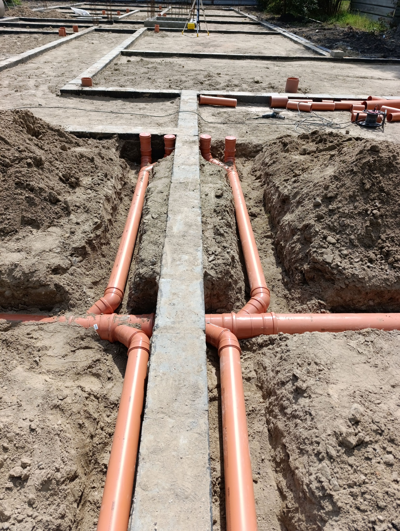 Pomarańczowe rury kanalizacyjne ułożone w wykopie, widoczne fundamenty budynku w tle.