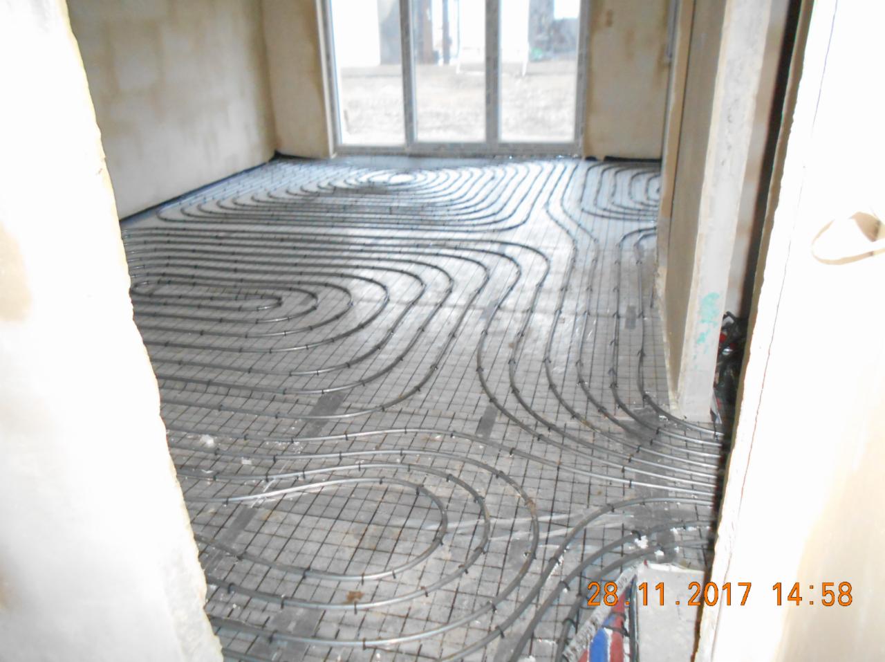 Równomiernie rozłożone rury ogrzewania podłogowego na siatce montażowej w surowym pomieszczeniu z oknami balkonowymi, przygotowane do zalania wylewką.
