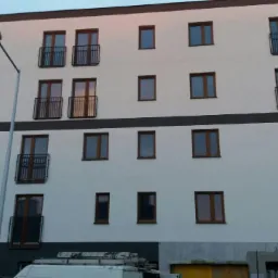 Nowoczesny budynek wielorodzinny z balkonami z czarnymi balustradami, widok z dołu na elewację z drewnianymi oknami, częściowo zasłonięty przez samochód dostawczy.