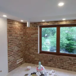 Pokój w trakcie wykańczania: ściana z cegły klinkierowej, brązowe okno, widoczne puszki, butelka wody i narzędzia glazurnicze na podłodze.
