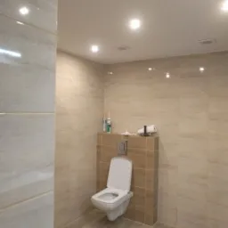 Nowoczesna łazienka z beżowymi płytkami imitującymi drewno, podwieszana toaleta z otwartą klapą na tle zabudowy z płytek, widoczne tuby silikonu na półce.