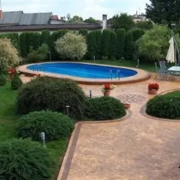 Owalny basen z błękitną wodą otoczony zielonym trawnikiem i dekoracyjnym, wzorzystym betonem, widok z góry na wypielęgnowany ogród z tujami, parasolem i meblami ogrodowymi