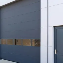 Szara brama garażowa segmentowa z przeszkleniami w dolnej części, obok szare drzwi wejściowe do budynku o białej elewacji z paneli.