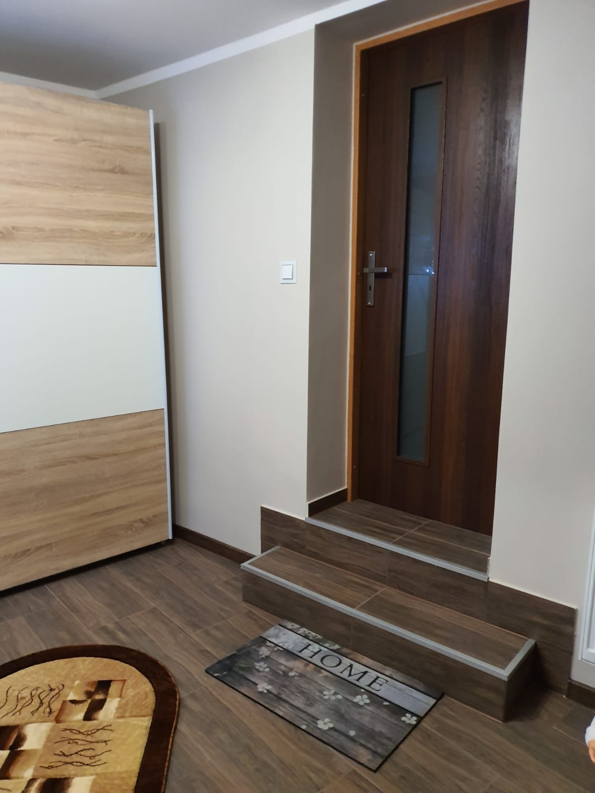 Wnętrze mieszkania z drewnopodobną podłogą i schodkami wyłożonymi płytkami, prowadzącymi do ciemnych drzwi z szybą; obok szafa z przesuwnymi drzwiami w odcieniach bieli i drewna.