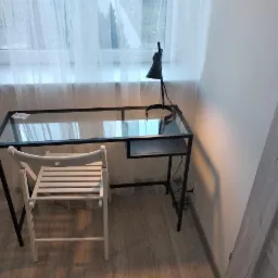 Szklane biurko z czarną metalową ramą i białe składane krzesło na tle okna z białymi zasłonami, minimalistyczny design.