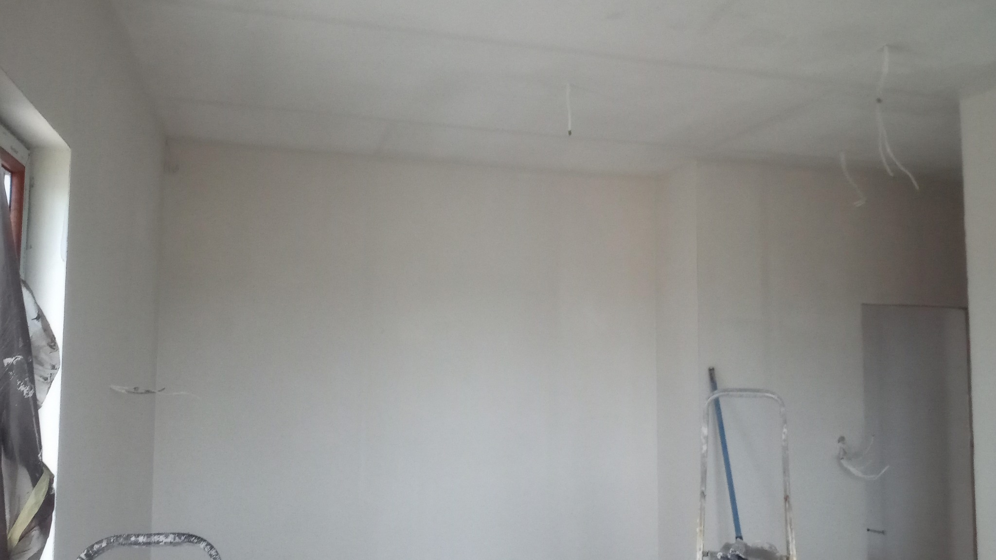 Świeżo pomalowane na biało wnętrze z wystającymi kablami z sufitu, drabina oparta o ścianę i zasłonięte okno po lewej stronie.