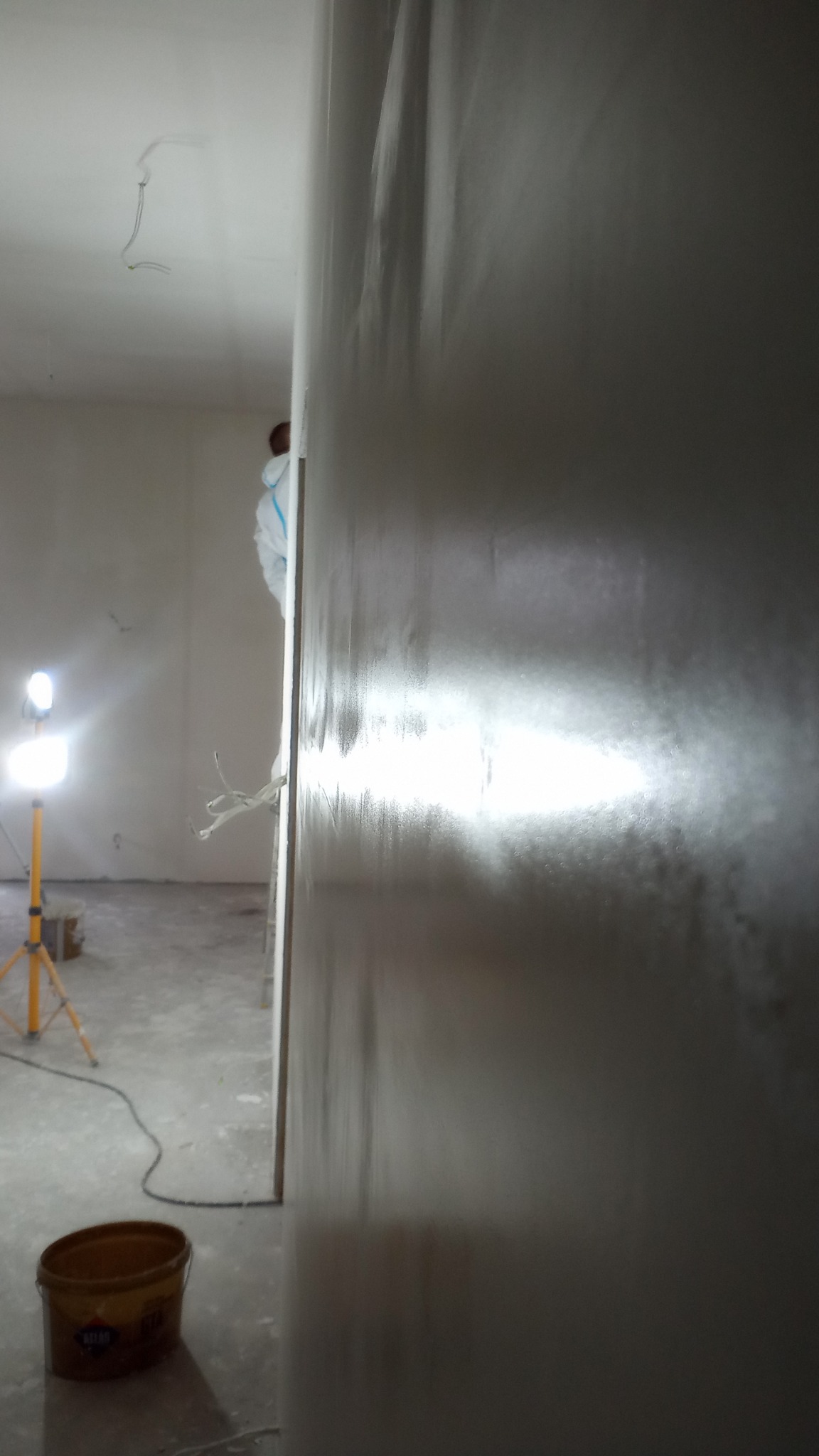 Świeżo nałożona, mokra gładź na ścianie, oświetlona lampą budowlaną, widoczny pracownik w kombinezonie ochronnym i wiadro z zaprawą.