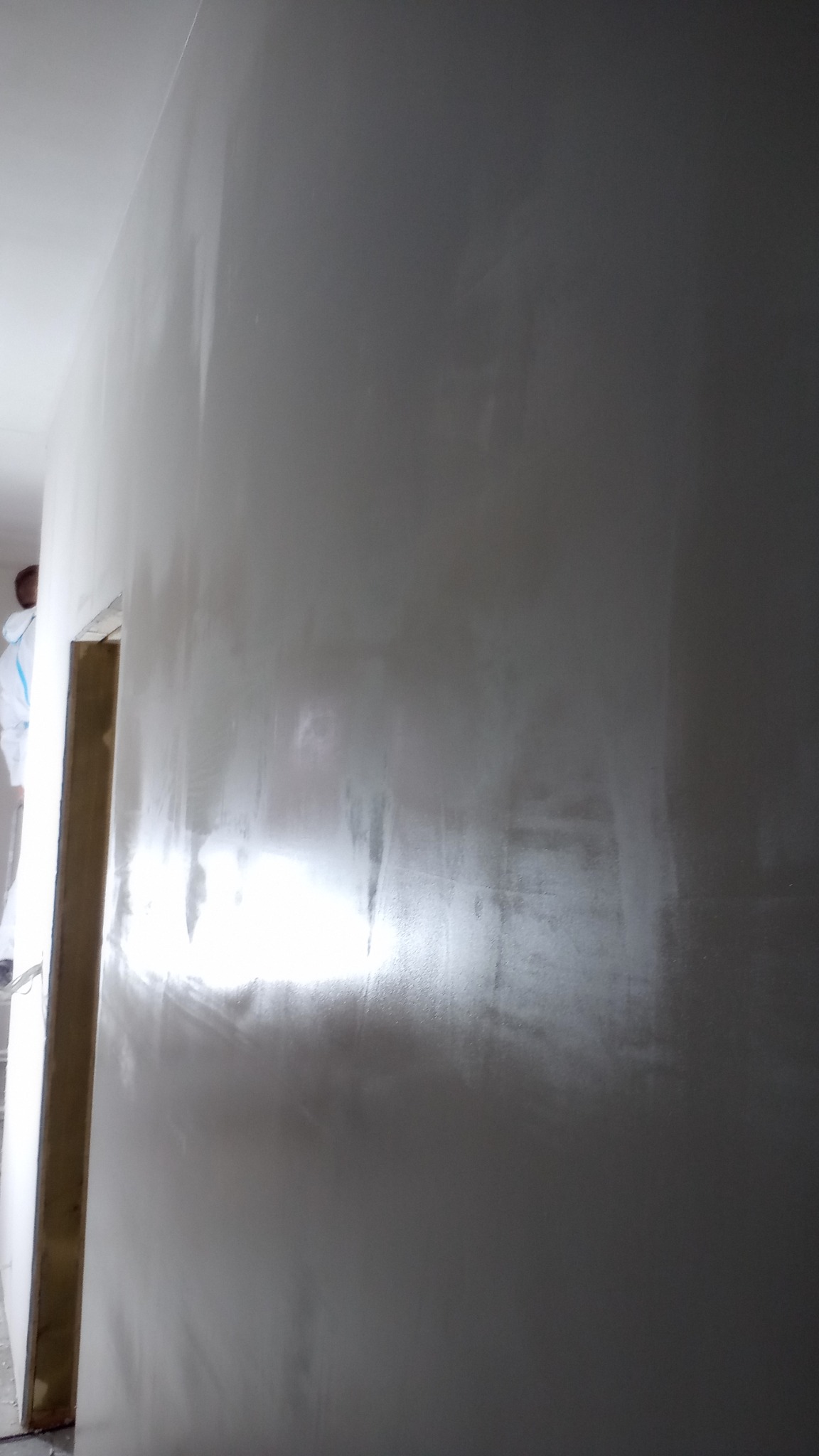 Świeżo położona, błyszcząca gładź na ścianie, widoczny fragment ościeżnicy i pracownik w kombinezonie malarskim w tle.