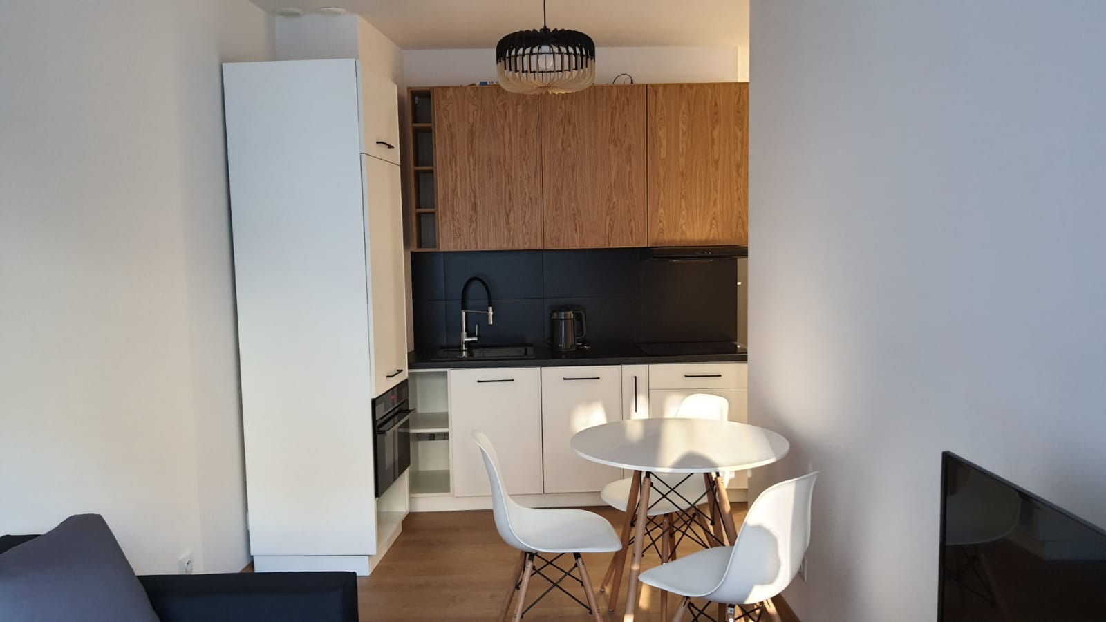 Nowoczesna, jasna kuchnia z drewnianymi szafkami wiszącymi i czarnym panelem nad blatem, okrągły biały stół z trzema białymi krzesłami w stylu skandynawskim.