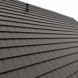 Ciemnoszary dach z blachy na rąbek stojący z kominkiem wentylacyjnym, widoczny z dołu pod kątem, na tle pochmurnego nieba.