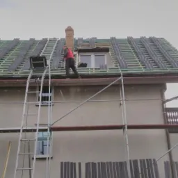 Pracownik w czerwonej bluzie na rusztowaniu układa dachówkę na dachu domu z ceglanym kominem, widoczny podnośnik do dachówki.