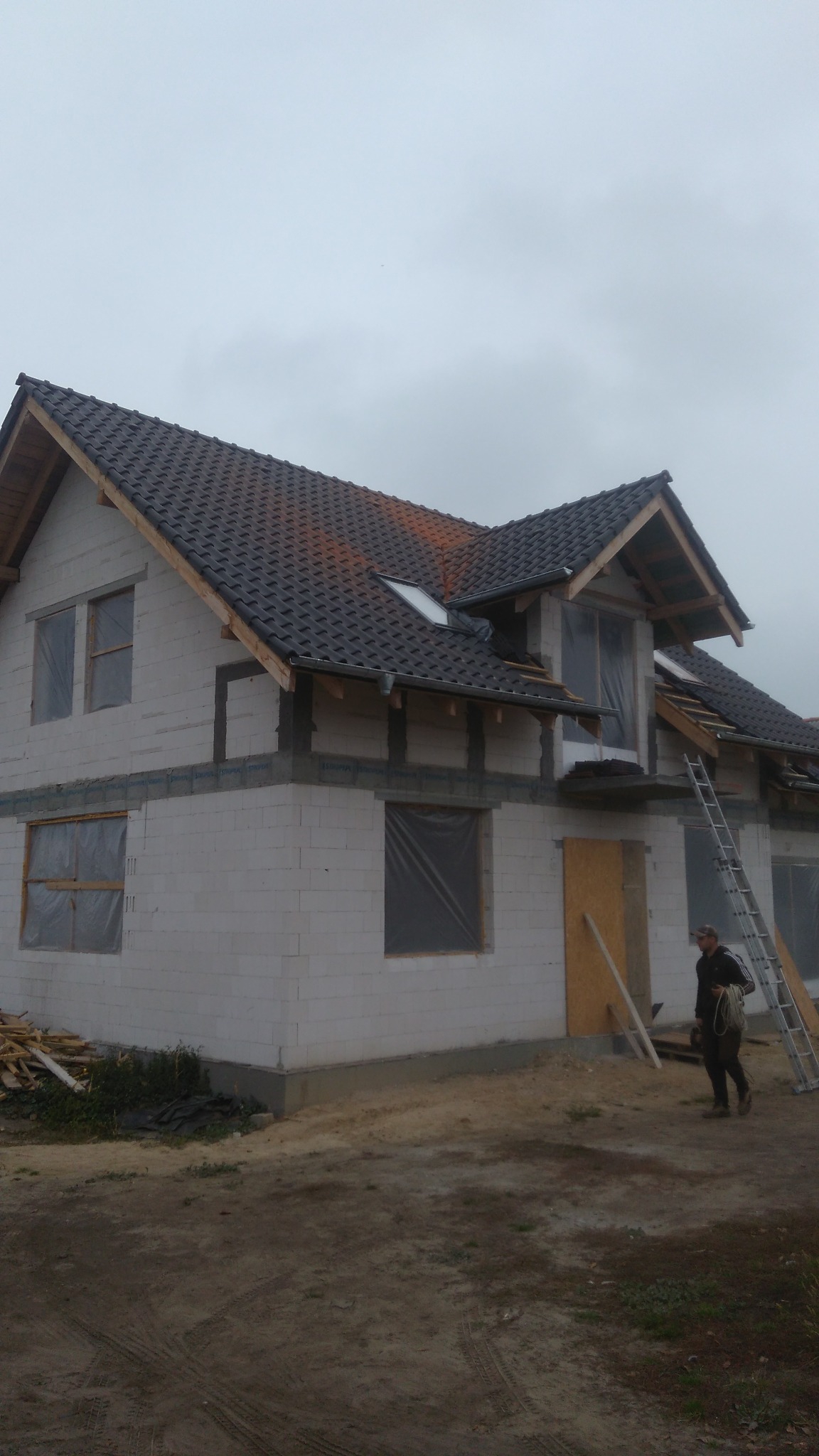 Dom w trakcie budowy z częściowo pokrytym dachem z ciemnej dachówki, widoczna więźba dachowa, okna zabezpieczone folią, drabina oparta o ścianę, na pierwszym planie mężczyzna z liną.