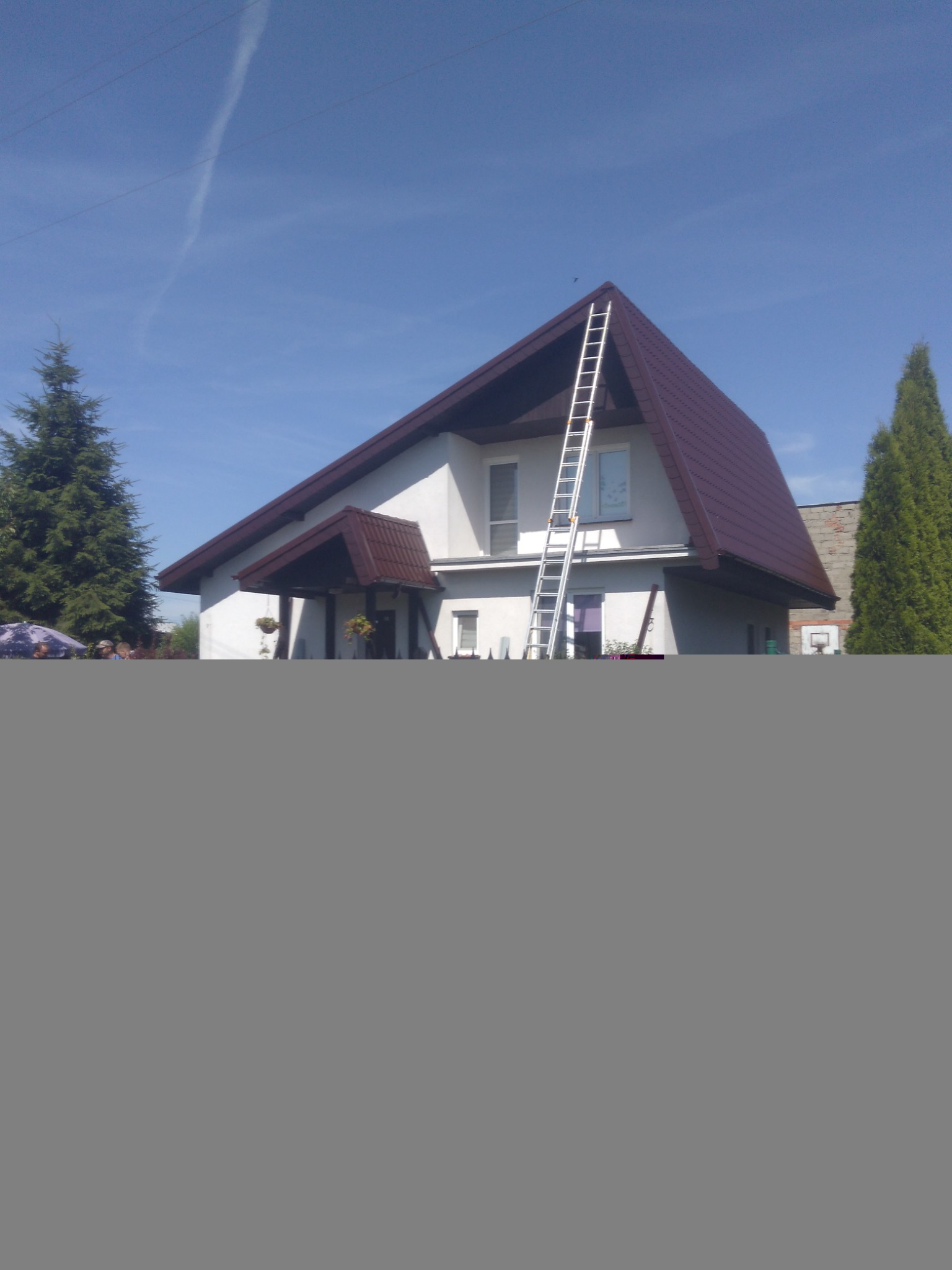 Dom jednorodzinny z dwuspadowym dachem z dachówki w kolorze brązowym, z drabiną opartą o krawędź dachu, widok z poziomu gruntu, błękitne niebo.