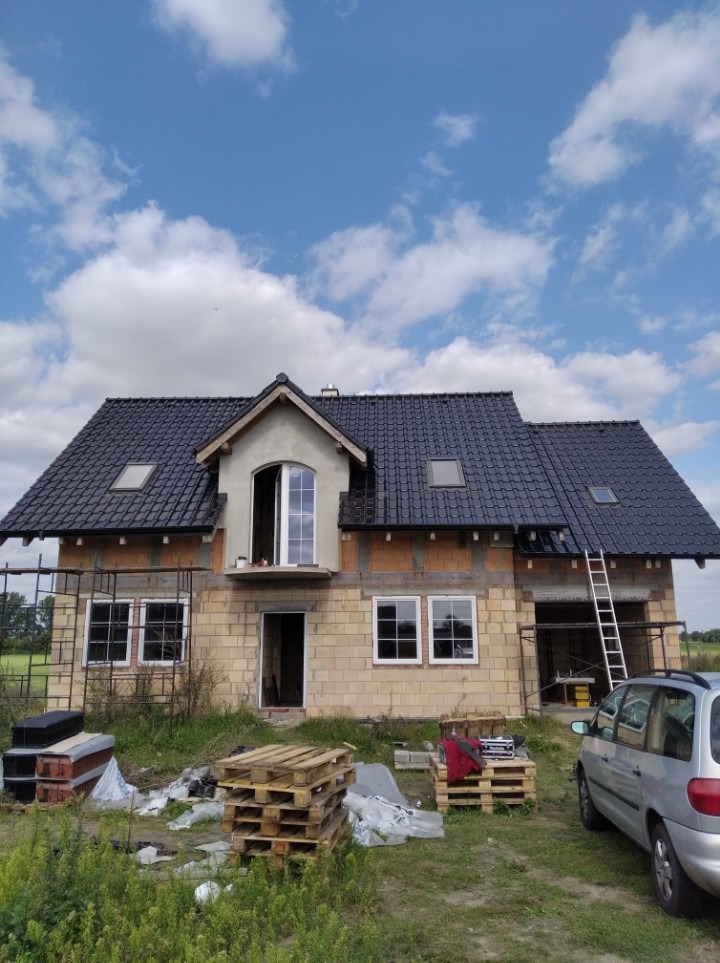 Dom w trakcie budowy z częściowo ukończonym dachem z czarnej dachówki, widoczne okna dachowe, ściany z pustaków, rusztowanie i drabina oparta o budynek, na pierwszym planie palety i samochód.