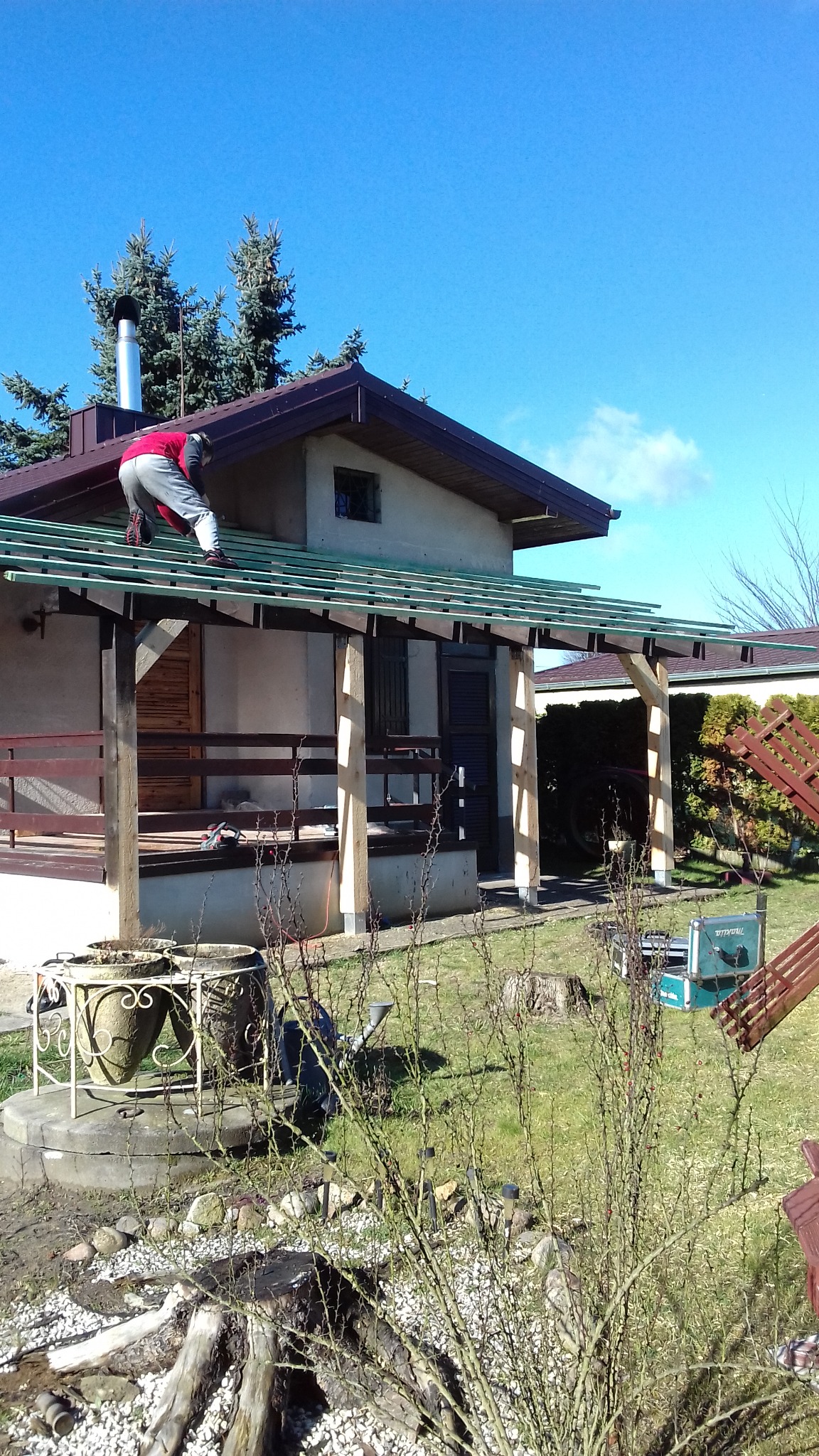 Pracownik w czerwonej bluzie montuje konstrukcję drewnianą na zadaszeniu werandy domu jednorodzinnego w słoneczny dzień.