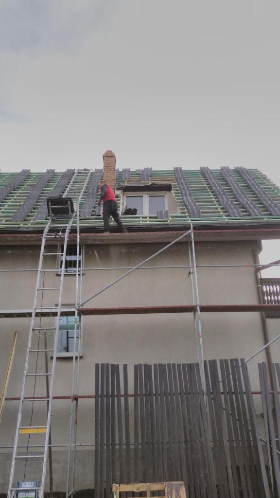 Pracownik w czerwonej bluzie na rusztowaniu układa dachówkę na dachu domu z ceglanym kominem, widoczny podnośnik do dachówki.