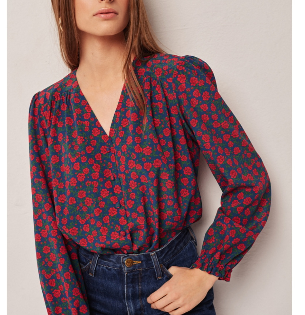 Kobieta w bluzce z długim rękawem w drobne czerwone róże na granatowym tle, z głębokim dekoltem w serek i dżinsach.