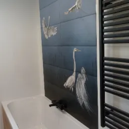 Nowoczesna łazienka z wanną obudowaną drewnopodobną płytą, ściana wyłożona płytkami z motywem czapli, czarna bateria wannowa i grzejnik drabinkowy.