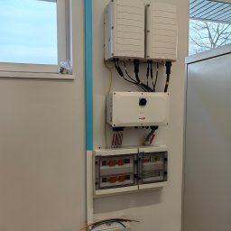 Zilmaks Instalację Zbigniew Zaręba - Biała ściana z zamontowanymi elementami instalacji fotowoltaicznej: dwa inwertery, skrzynka rozdzielcza i moduł SolarEdge. Widoczne okno i rura w kolorze błękitnym.