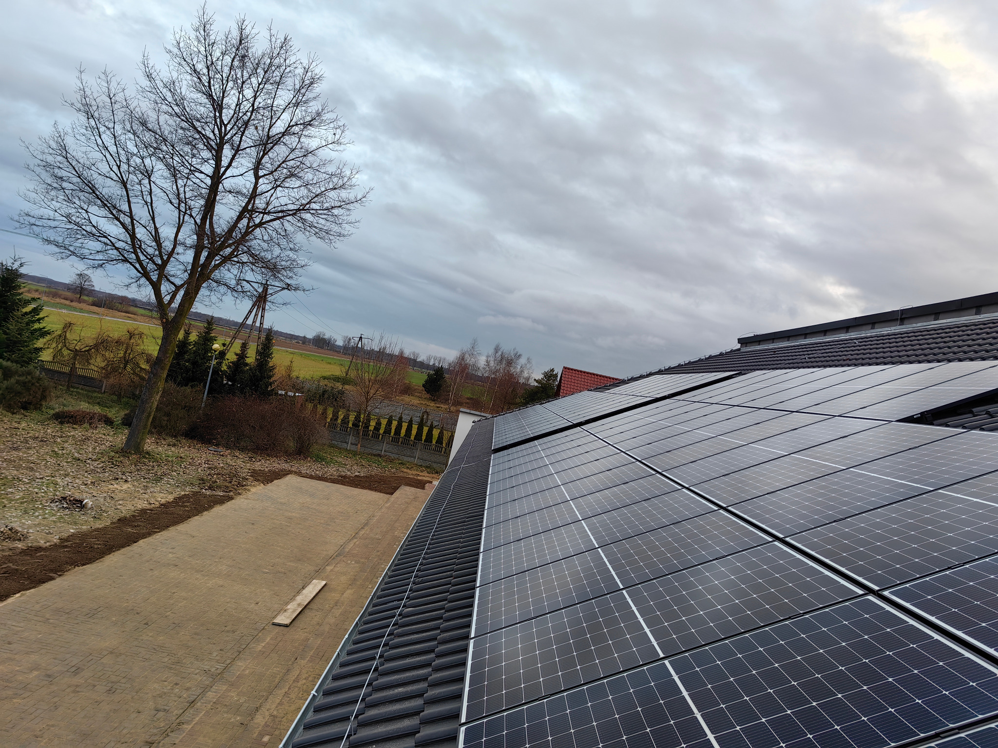 Instalacja fotowoltaiczna na dachu domu jednorodzinnego, widok z góry, z drzewem bez liści i pochmurnym niebem w tle. Panele w rzędach, czyste i równe.