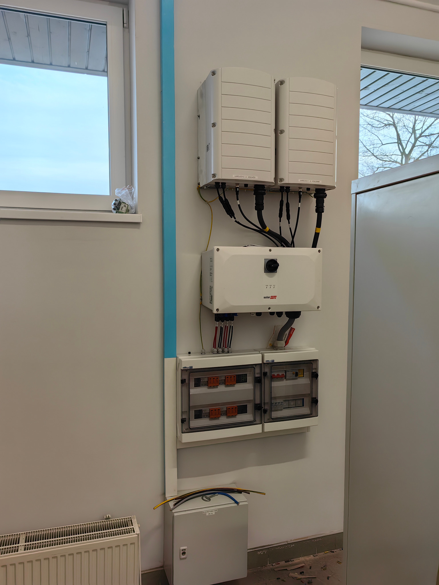 Biała ściana z zamontowanymi elementami instalacji fotowoltaicznej: dwa inwertery, skrzynka rozdzielcza i moduł SolarEdge. Widoczne okno i rura w kolorze błękitnym.
